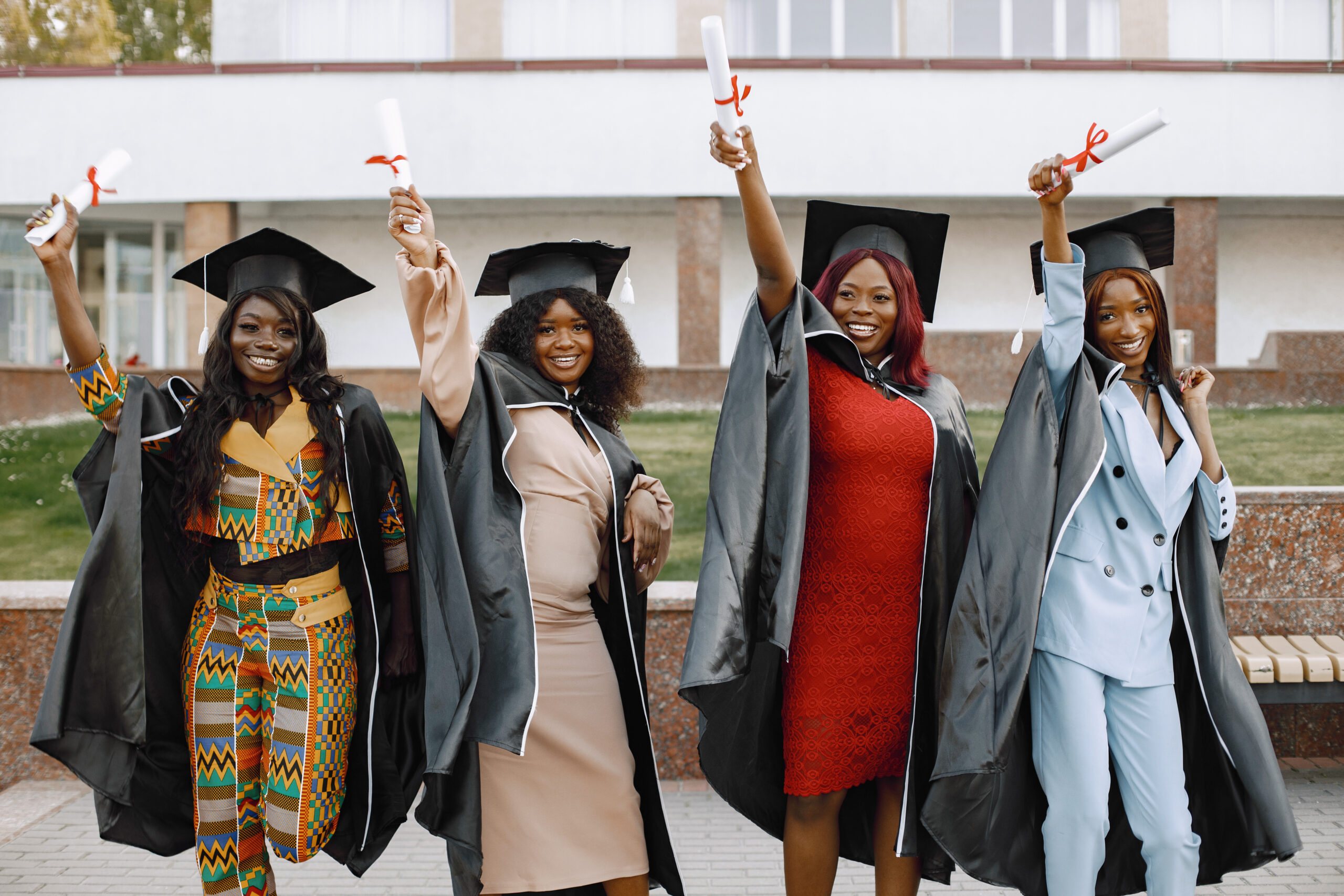 Group of young afro american female student dressed in black graduation gown. Campus as a background. Girls cheerfully smiling with arms up, holding diploma.